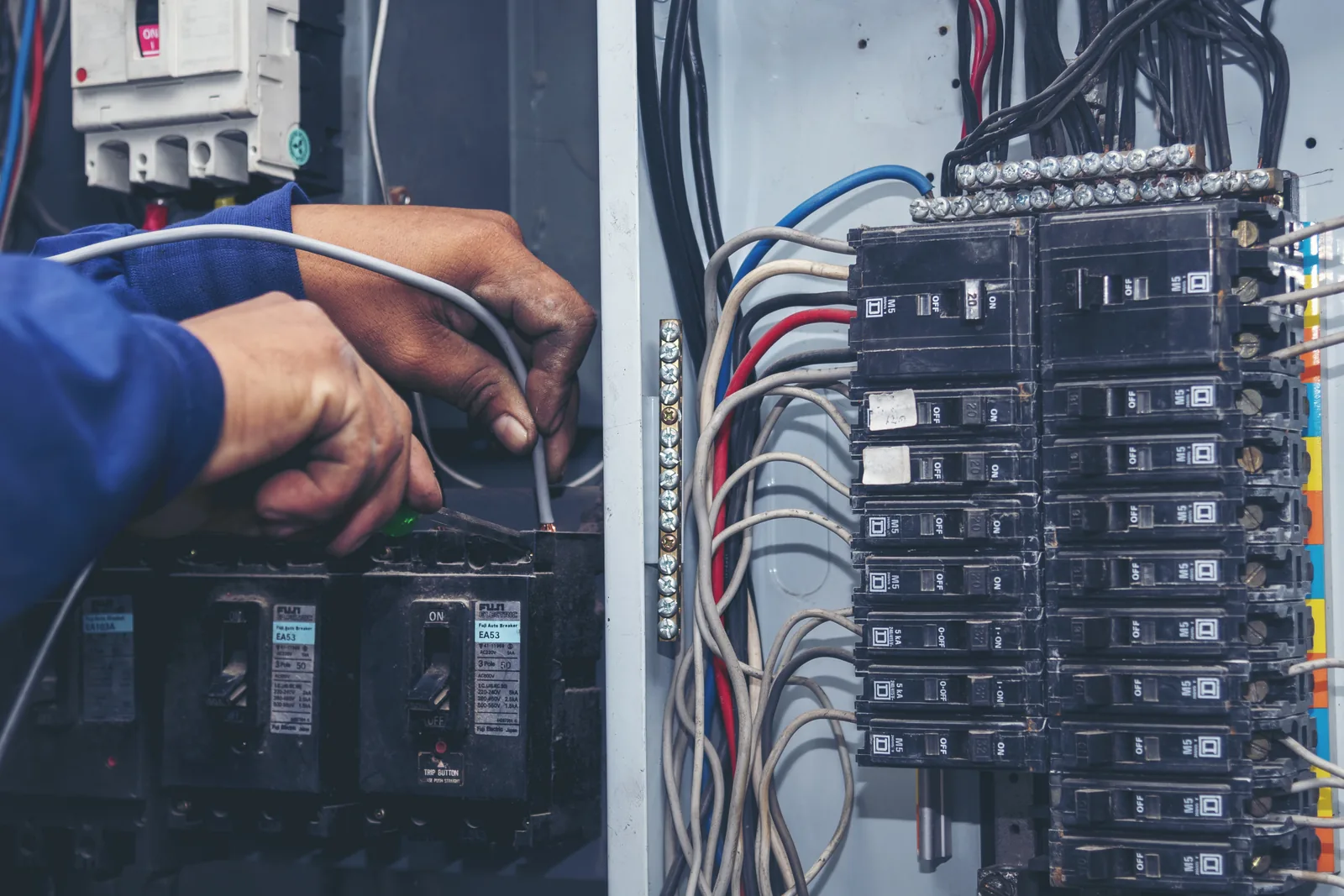 An electrician working inside an open switchboard, testing circuit breaker wiring